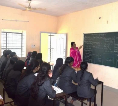 A teacher in a bright pink dress standing at a blackboard explaining a lesson to a classroom of seated students.