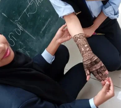 A close-up of a student applying an intricate henna (mehndi) design on another student's hand.